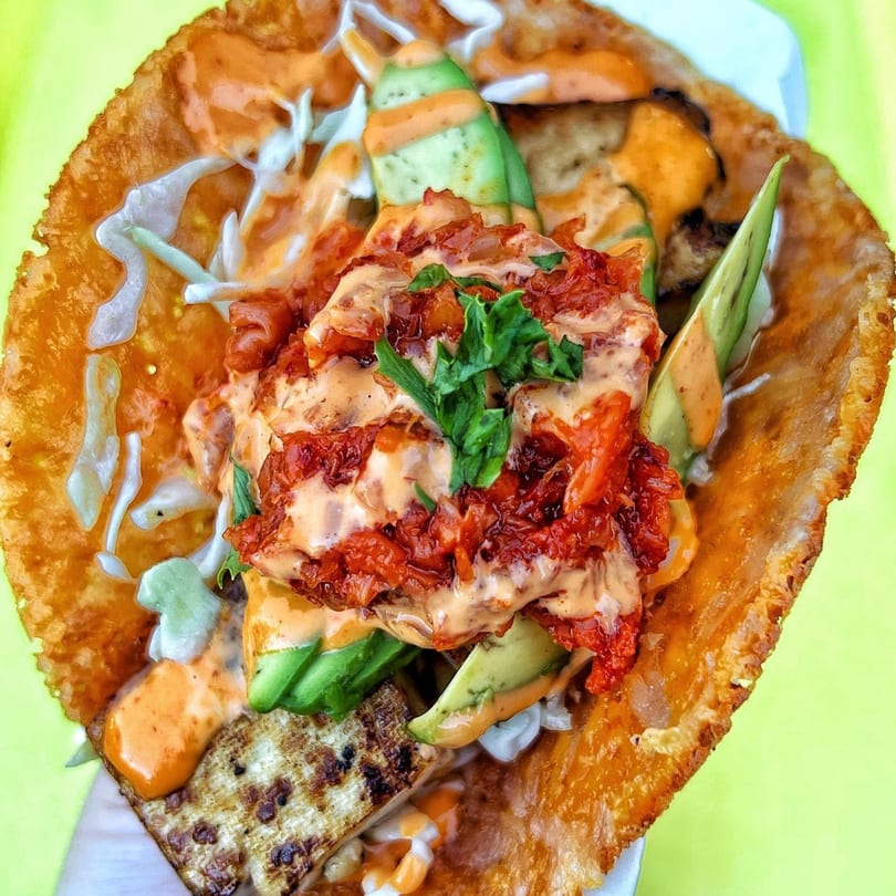 A crispy Korean-Mexican taco with kimchi, avocado, and creamy sauces on a bright green background.
