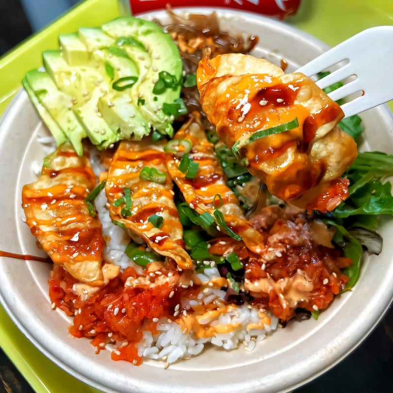 A fork lifts a Korean dumpling from a bowl of rice, avocado, kimchi, and other toppings.
